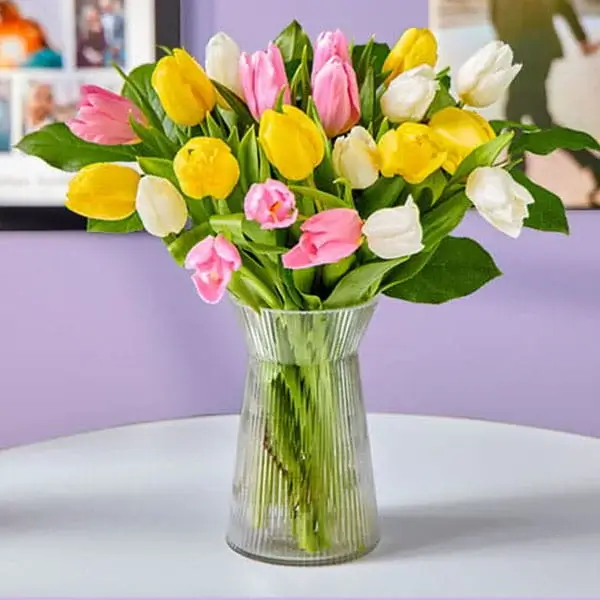 A clear ribbed glass vase filled with yellow, pink, and white tulips, arranged in a full bouquet on a white table against a soft purple background.